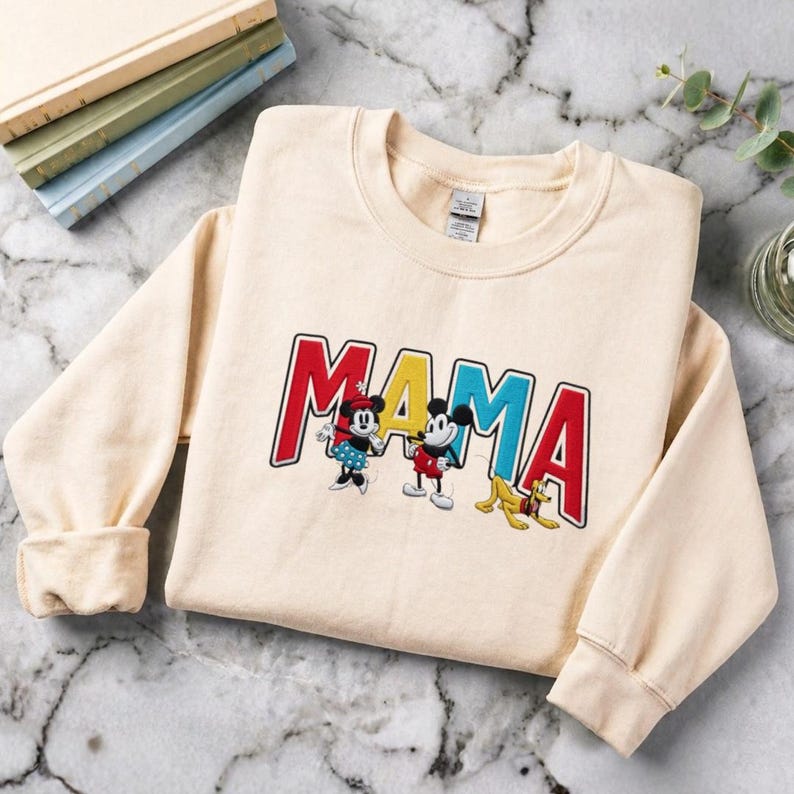 Puede incluir: Sudadera color crema con la palabra "MAMA" bordada en letras rojas, azules y amarillas. La sudadera presenta im&aacute;genes bordadas de Minnie Mouse, Mickey Mouse y Pluto. La sudadera tiene mangas largas y cuello redondo.