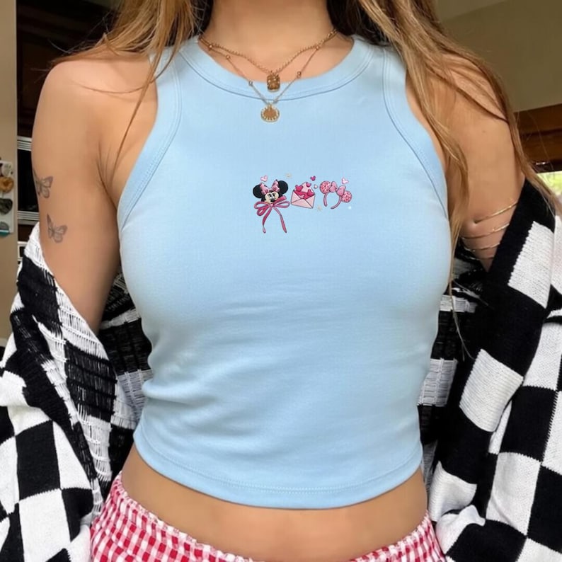 May include: Pale blue vest top with a round neck. The top has an embroidered design of Minnie Mouse, a letter, and a headband. The model is wearing a black and white checkered cardigan and a red and white checkered skirt.