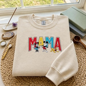 Puede incluir: Una sudadera de color crema con la palabra "MAMA" en letras rojas, amarillas y azules. La sudadera presenta los personajes de dibujos animados Minnie Mouse, Mickey Mouse y Pluto. La sudadera est&aacute; hecha de un material suave.