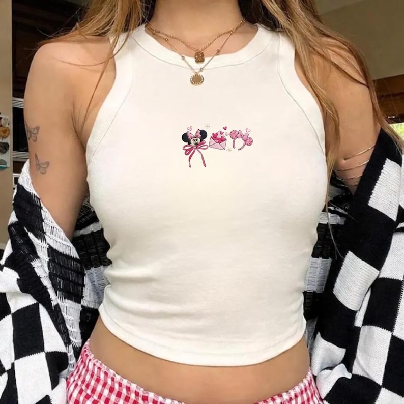May include: A white crop top with a scoop neck and a small Minnie Mouse graphic in pink and red. The graphic includes a bow, envelope, and heart-shaped details. The top is sleeveless and made of a soft material.