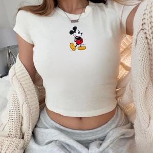 Puede incluir: Top corto de color crema con mangas cortas y un gr&aacute;fico cl&aacute;sico de Mickey Mouse. El personaje de dibujos animados est&aacute; representado con sus ic&oacute;nicos pantalones cortos rojos, zapatos amarillos y guantes negros. La parte superior est&aacute; hecha de un material suave.