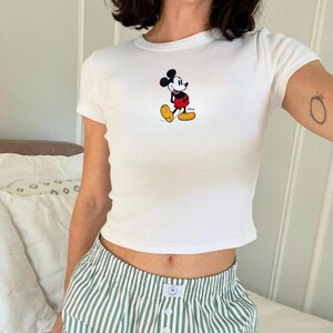 Puede incluir: Camiseta blanca de manga corta con un gr&aacute;fico de Mickey Mouse en negro, rojo, amarillo y blanco. La camiseta se combina con pantalones cortos a rayas verdes y blancas. Dise&ntilde;o cl&aacute;sico de personaje de dibujos animados.