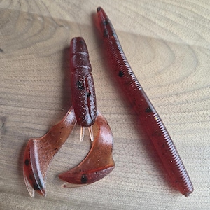 May include: Two red and brown fishing lures. One lure is shaped like a crawfish with two claws and a segmented body. The other lure is a long, slender worm with a ribbed body. Both lures have black speckles.