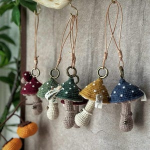 May include: A collection of handmade crochet mushroom ornaments in various colors, including burgundy, green, yellow, and blue. Each mushroom has white polka dots on the cap and a beige stem. They are suspended from a wooden branch with metal hooks and copper-colored wire.