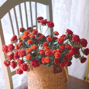 Könnte beinhalten: Ein Weidenkorb gefüllt mit handgefertigten roten Häkelrosen. Die Rosen haben grüne Blätter und Stiele. Der Korb steht auf einem Holzstuhl. Der Hintergrund ist ein weißer Vorhang.