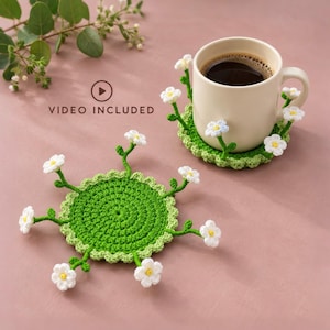 May include: Two round crochet coasters in bright green with a scalloped edge, each featuring several small white flowers with yellow centres attached to thin green stems extending outward. One coaster supports a beige ceramic mug filled with coffee.