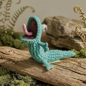 Może przedstawiać: Szydełkowe stworzenie w kolorze morskim z otwartą paszczą, różowym językiem i białym okiem. Ręcznie robione amigurumi jest ułożone na kawałku drewna, z mchem i małymi kamieniami w tle. Stworzenie ma małe ramiona i długi ogon.