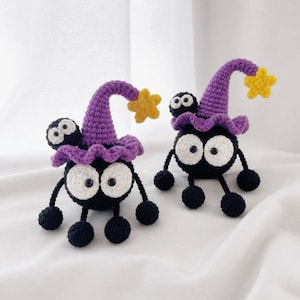 May include: Two round black crochet creatures with large white eyes and black pupils. Each figure wears a purple pointed hat with a yellow star at the tip and a small black ball-like attachment on the side. They have thin black limbs with round feet.