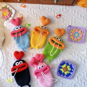 Puede incluir: Una colecci&oacute;n de art&iacute;culos de ganchillo coloridos y hechos a mano. La imagen presenta varias fundas de tel&eacute;fono caprichosas en azul, amarillo, verde, negro y rosa, cada una con ojos saltones, una boca roja o rosa y un acento de coraz&oacute;n. Tambi&eacute;n se incluyen cuadrados de ganchillo.