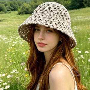 May include: A beige crochet sun hat with a wide brim. The hat features an open, geometric pattern. The image is set in a field of wildflowers with a green, mountainous background. The hat is a stylish accessory for sunny days.