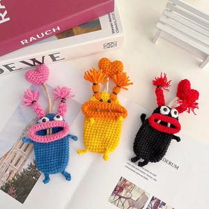Puede incluir: Tres figuras hechas a mano de crochet con forma de monstruo en azul, amarillo y negro. Cada una tiene una boca muy abierta, ojos, brazos y piernas. La figura azul tiene un coraz&oacute;n rosa y pelo rosa. La figura amarilla tiene pelo naranja y un coraz&oacute;n. La figura negra tiene pelo rojo y un coraz&oacute;n.