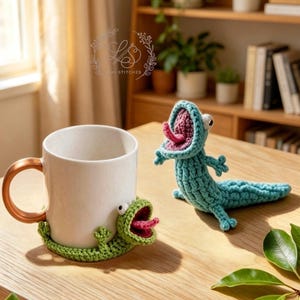 May include: A white ceramic mug with a copper-colored handle, accompanied by a green crocheted lizard coaster and a matching turquoise lizard figure. The lizard's mouth is open, revealing a pink tongue. The items are on a wooden surface.
