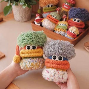 May include: Handmade crochet dolls with large googly eyes, featuring fuzzy hair and colorful details. One doll has green hair and a yellow and orange face, while the other has gray hair and an orange face. Other dolls are in a wooden tray.