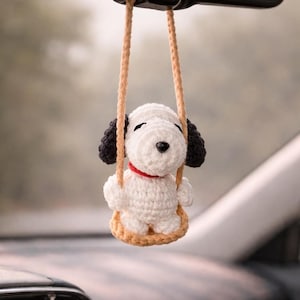 Puede incluir: Un adorno de coche de perro Snoopy tejido a ganchillo colgando del espejo retrovisor de un coche. El perro blanco tiene orejas negras y un collar rojo, sentado en un columpio beige. El fondo está borroso, sugiriendo el interior de un coche.