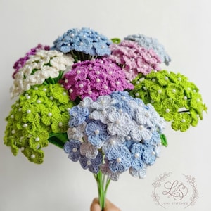 Puede incluir: Un ramo de flores de ganchillo hechas a mano en varios colores, incluyendo azul, morado, verde y blanco. Cada flor está adornada con pequeñas cuentas blancas. Las flores se sujetan con tallos verdes.