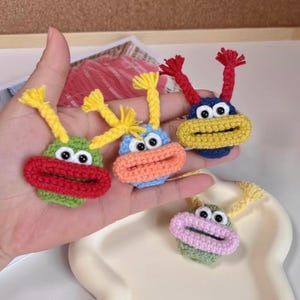 May include: A collection of handmade crochet monster face ornaments in various colors. Each ornament features a round body with a wide mouth, googly eyes, and yarn antennae. Colors include green, red, blue, yellow, and pink.