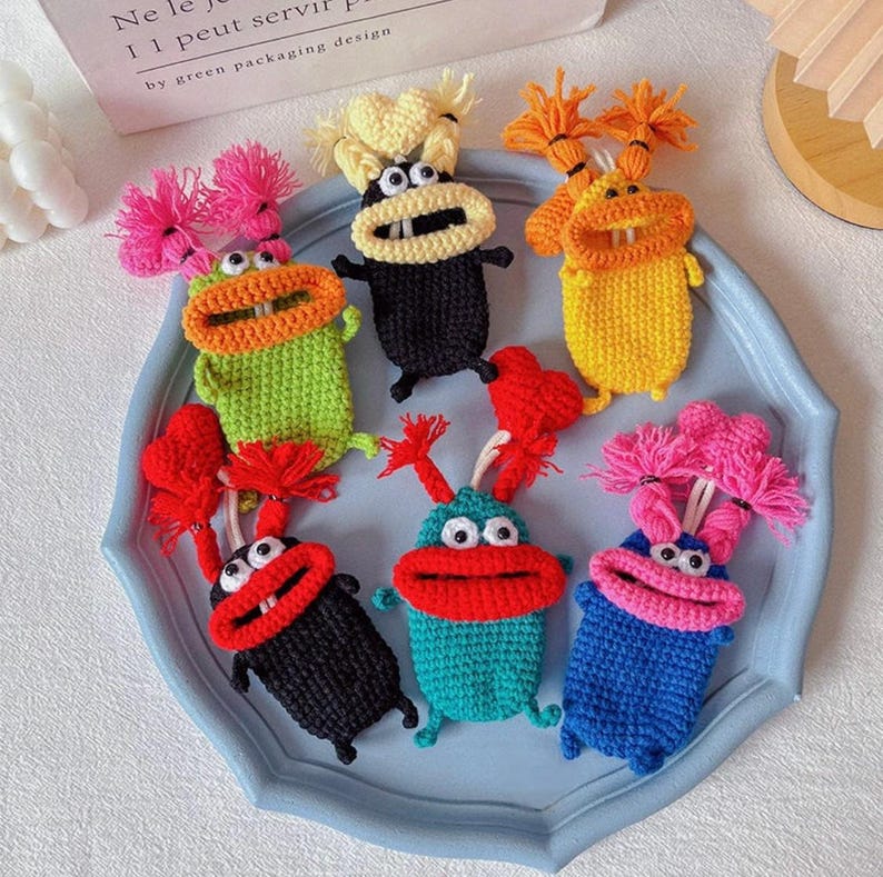 Puede incluir: Una colecci&oacute;n de figuras de monstruos de ganchillo hechas a mano y coloridas. Cada figura tiene una combinaci&oacute;n de colores &uacute;nica, que incluye verde, negro, amarillo, azul y rojo, con bocas grandes y cabello caprichoso. Est&aacute;n dispuestas en una bandeja azul claro.