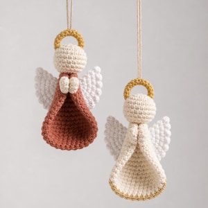 Puede incluir: Dos adornos colgantes de &aacute;ngeles tejidos a ganchillo con cabezas color crema, halos dorados y alas blancas. Un &aacute;ngel tiene un vestido color terracota, mientras que el otro tiene un vestido color crema con borde dorado, suspendidos por cordel.