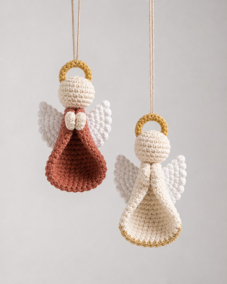 Puede incluir: Dos adornos colgantes de &aacute;ngeles tejidos a ganchillo con cabezas color crema, halos dorados y alas blancas. Un &aacute;ngel tiene un vestido color terracota, mientras que el otro tiene un vestido color crema con borde dorado, suspendidos por cordel.