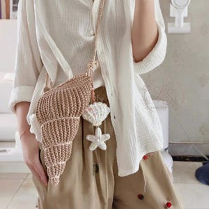 May include: A beige crochet seashell-shaped crossbody bag with a matching strap. The bag is adorned with a white seashell and starfish charm. The bag is part of a beach-themed collection.