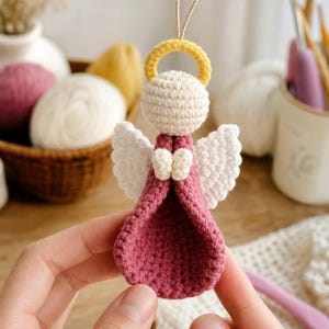 Puede incluir: Un adorno de &aacute;ngel tejido a ganchillo con un vestido rosa empolvado, cabeza color crema, alas blancas y un halo amarillo. La figura incluye un peque&ntilde;o lazo de cuerda beige en la parte superior para colgar, con una construcci&oacute;n de hilo texturizado.