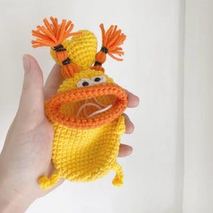Puede incluir: Una bolsa de ganchillo amarilla hecha a mano con forma de personaje de dibujos animados con pelo naranja y boca abierta. La bolsa tiene ojos blancos y labios naranjas. El personaje tiene dos piernas peque&ntilde;as. La bolsa se sostiene en una mano.