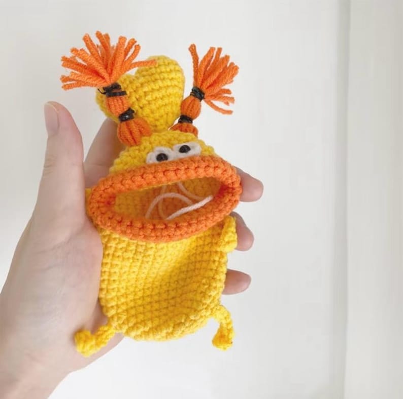 Puede incluir: Una bolsa de ganchillo amarilla hecha a mano con forma de personaje de dibujos animados con pelo naranja y boca abierta. La bolsa tiene ojos blancos y labios naranjas. El personaje tiene dos piernas peque&ntilde;as. La bolsa se sostiene en una mano.