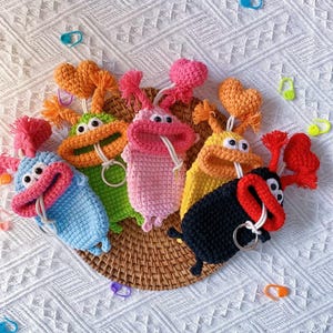 Puede incluir: Una colecci&oacute;n de llaveros con forma de monstruo de ganchillo de colores. Cada llavero presenta un color &uacute;nico, incluyendo azul, verde, rosa, amarillo y negro, con ojos saltones y un detalle de coraz&oacute;n o pomp&oacute;n. Est&aacute;n dispuestos sobre una cesta tejida.