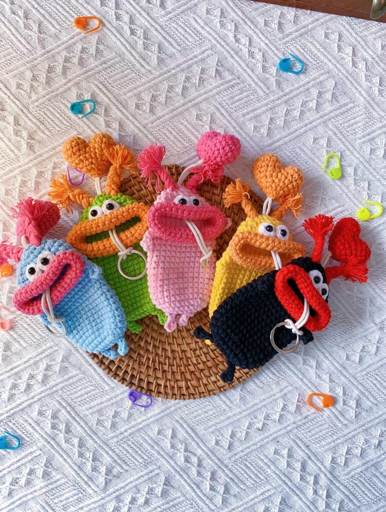 Puede incluir: Una colecci&oacute;n de llaveros con forma de monstruo de ganchillo de colores. Cada llavero presenta un color &uacute;nico, incluyendo azul, verde, rosa, amarillo y negro, con ojos saltones y un detalle de coraz&oacute;n o pomp&oacute;n. Est&aacute;n dispuestos sobre una cesta tejida.