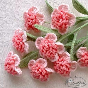 Könnte beinhalten: Handgefertigte Häkelblumen in Rosa und Weiß mit grünen Blättern. Die Blumen haben eine strukturierte Optik. Die Anordnung befindet sich vor einem weißen Spitzengrund, wodurch eine zarte und künstlerische Darstellung entsteht.