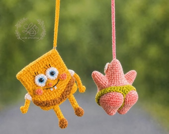 May include: Two hanging crochet ornaments suspended by strings. One is a yellow rectangular character with blue eyes and a brown base. The other is a pink starfish character wearing green trunks. Both are crafted with yarn and feature detailed stitching.