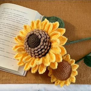 May include: Handmade crocheted sunflowers with yellow petals and brown centres, green stems and leaves, lie on a textured brown surface next to an open book. The flowers are detailed and vibrant.