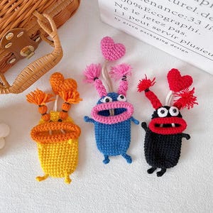 Puede incluir: Tres llaveros de monstruos hechos a mano con ganchillo en amarillo, azul y negro. Cada uno tiene una boca abierta, ojos saltones y pelo de hilo. Los monstruos azules y negros tienen corazones rosas y rojos, respectivamente. El monstruo amarillo tiene pelo naranja.