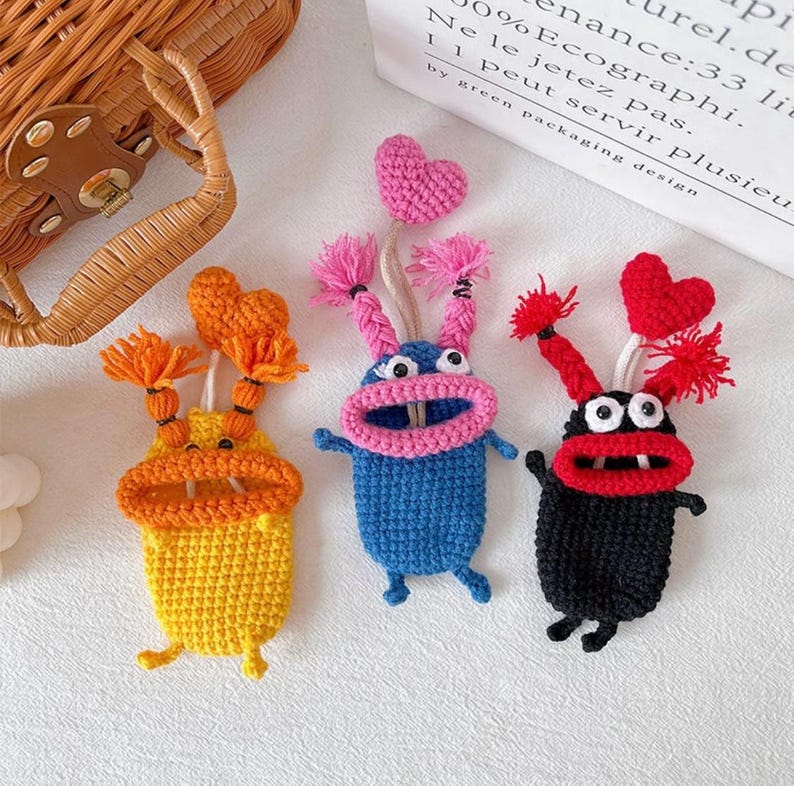 Puede incluir: Tres llaveros de monstruos hechos a mano con ganchillo en amarillo, azul y negro. Cada uno tiene una boca abierta, ojos saltones y pelo de hilo. Los monstruos azules y negros tienen corazones rosas y rojos, respectivamente. El monstruo amarillo tiene pelo naranja.
