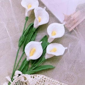 Könnte beinhalten: Ein handgefertigter Häkelstrauß mit Calla-Lilien. Die Blüten sind weiß mit gelben Zentren, grünen Stielen und grünen Blättern. Der Strauß ist mit einem cremefarbenen Spitzenband gebunden.