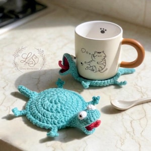 May include: Two handmade crochet coasters in the shape of a blue monster with a red tongue. One coaster supports a beige mug with a brown handle and a cat illustration. A white spoon rests nearby. The coasters are handmade.