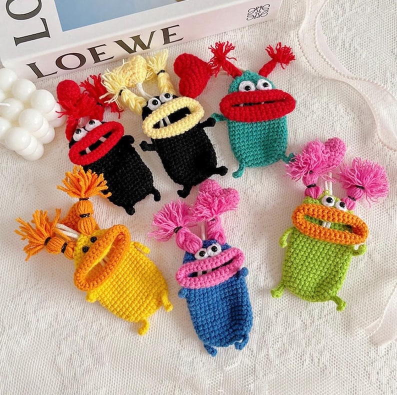Puede incluir: Una colecci&oacute;n de llaveros con forma de monstruo de ganchillo de colores. Cada llavero presenta una combinaci&oacute;n de colores &uacute;nica, incluyendo rojo, negro, amarillo, azul, verde y rosa. Tienen bocas grandes, ojos saltones y detalles de pomp&oacute;n. La palabra "LOEWE" es visible en el fondo.