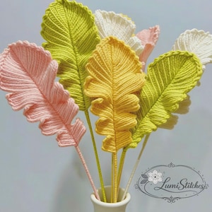 Puede incluir: Un ramo de hojas hechas a mano con hilo en un jarrón blanco. Las hojas son de varios colores, incluyendo rosa, amarillo, verde y blanco. Los tallos son delgados y verdes. Las hojas tienen un diseño detallado y texturizado. El jarrón es blanco y cilíndrico.