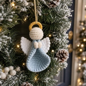 Puede incluir: Un adorno de &aacute;ngel tejido a ganchillo con vestido azul claro, alas blancas, cabeza color crema y halo dorado. La figura tiene peque&ntilde;as manos juntas frente al cuerpo y est&aacute; suspendida por una cinta dorada fina para colgarla.