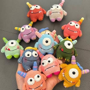 May include: A collection of colorful, crocheted monster toys. Each monster has one eye, a horn, and small arms and legs. The monsters are in various colors, including red, pink, blue, green, and yellow. They have smiling faces and are arranged on a gray surface.