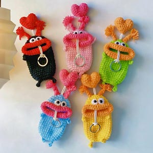 May include: A collection of five handmade crochet keychains in various colors. Each keychain features a whimsical creature design with large lips, googly eyes, and heart-shaped accents. The keychains include a metal ring for attaching keys.