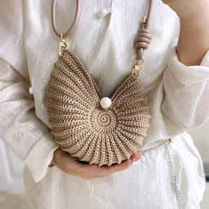 Könnte beinhalten: Eine beige, handgefertigte Häkeltasche mit Muscheldesign. Die Tasche hat eine runde, perlenartige Verzierung in der Mitte und einen langen, hellbraunen Lederriemen. Die Tasche wird von einer Person gehalten, die ein weißes Hemd trägt.