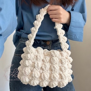 May include: Cream-colored, textured handbag with a unique, bubbly design. The bag has a rectangular shape with a matching strap. The bag is held by a person wearing a blue button-down shirt and blue jeans.