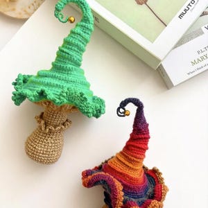 May include: Two handmade crochet mushroom figures with tall, curled caps and small gold bells at the tips. One mushroom has a bright green cap and tan stem, while the other features a multi-colored, gradient cap in red, orange, and purple tones.