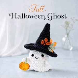 May include: A white crocheted ghost figurine wearing a black pointed witch hat adorned with orange and green leaves. The ghost has black eyes, a small smile, and pink cheeks, and is holding a small orange crocheted pumpkin in one hand.