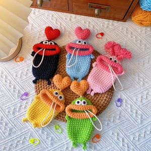 Puede incluir: Una colecci&oacute;n de monederos de ganchillo hechos a mano en varios colores, incluyendo negro, azul, rosa, amarillo y verde. Cada monedero presenta un dise&ntilde;o caprichoso de rana con ojos saltones, un coraz&oacute;n y un cierre de cord&oacute;n. Los monederos est&aacute;n colocados sobre una esterilla tejida.