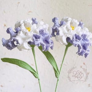 May include: Two handmade crochet flowers with white and lavender petals and yellow centers. The flowers have green stems and leaves. The background is a light, textured surface.