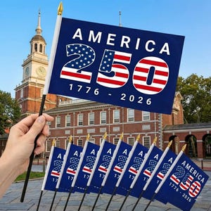 Puede incluir: Un conjunto de pequeñas banderas americanas con fondo azul, con el texto "AMERICA 250 1776-2026" en blanco y rojo. Las banderas se sostienen frente a un edificio histórico con una torre de reloj.
