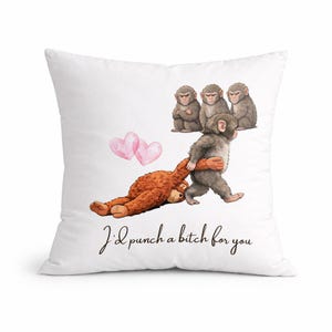 Puede incluir: Cojín blanco con una ilustración humorística de monos y un oso de peluche. La imagen presenta tres monos, dos corazones rosas y el texto "I'd punch a bitch for you" en escritura cursiva.