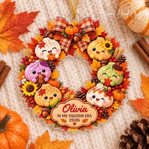 Op de afbeelding: Een cirkelvormig herfstornament met vijf lachende knoedel-figuren, omringd door herfstbladeren, dennenappels, eikels en kleine pompoenen. Een houten plaatje in het midden toont de tekst Olivia IN MY SQUISHY ERA 2026.